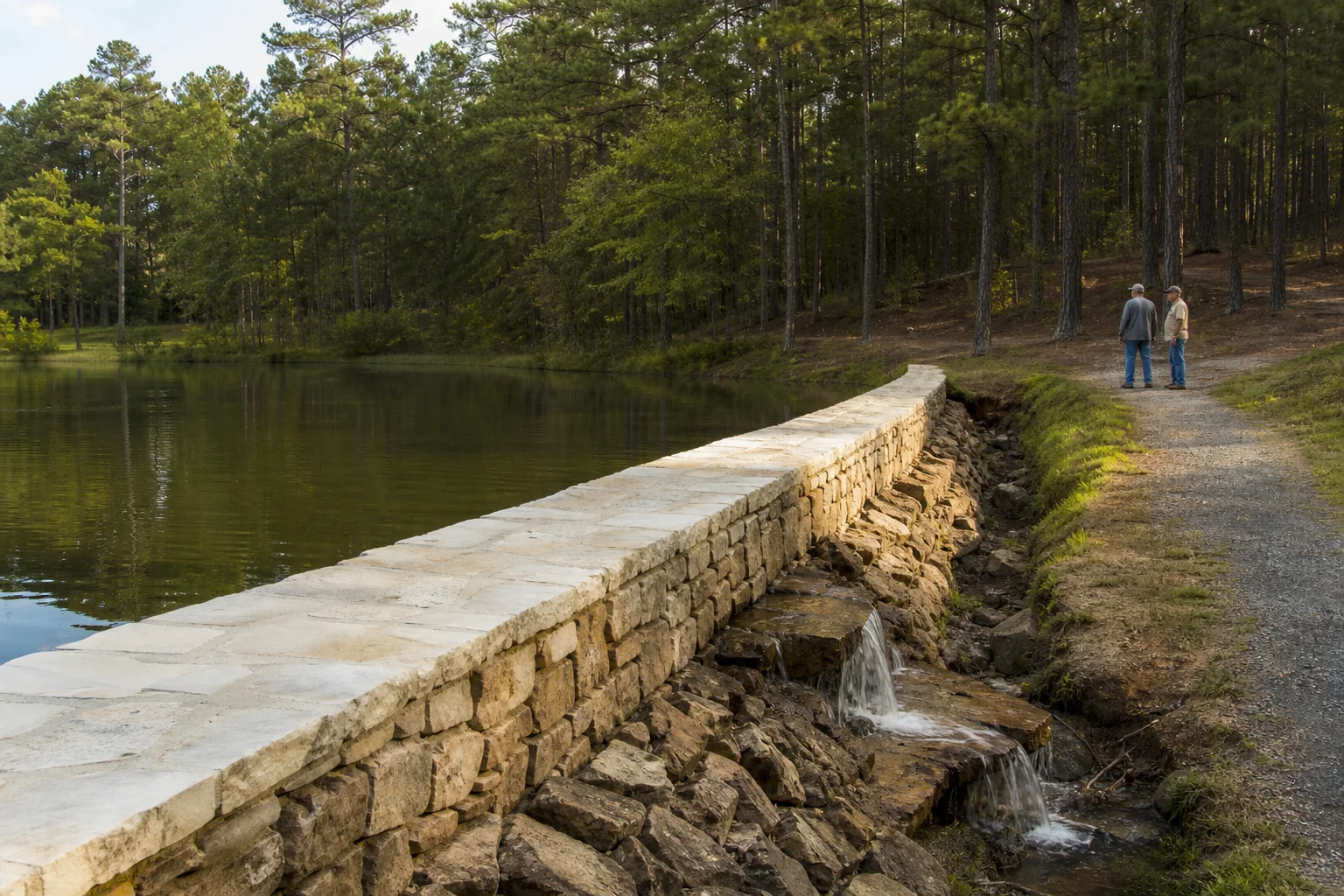 A small reinforced camp dam borders a calm lake among tall southern pine trees.