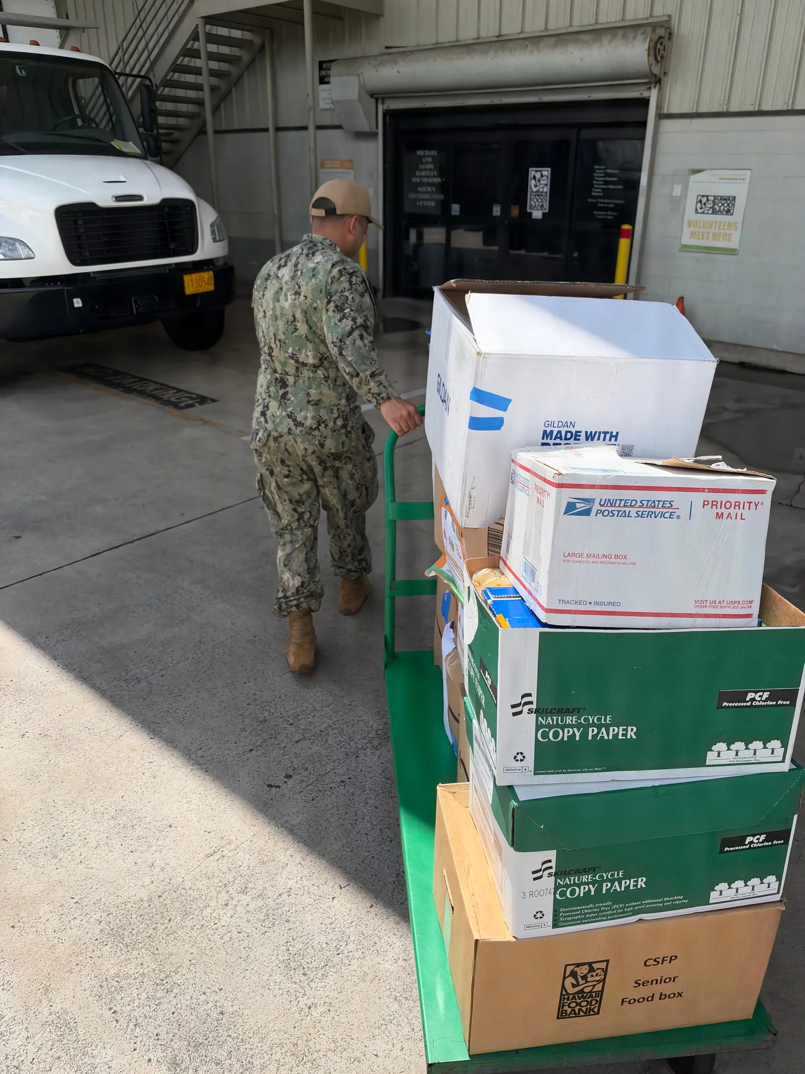 A Navy sailor guides a cart stacked with donated food boxes toward a Hawaiʻi Foodbank warehouse