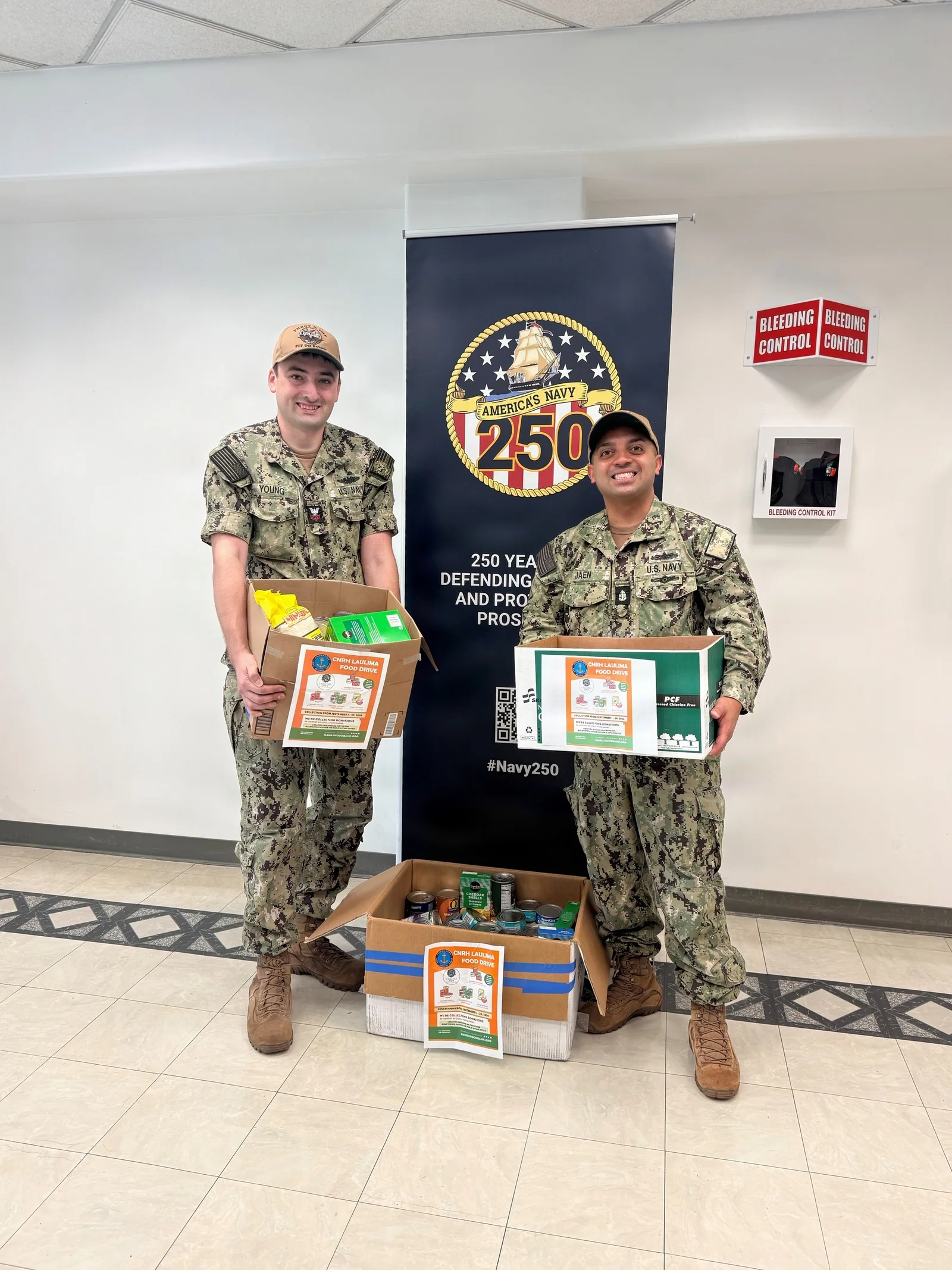 Two Navy sailors smile while holding boxes of donated pantry food at Joint Base Pearl Harbor-Hickam